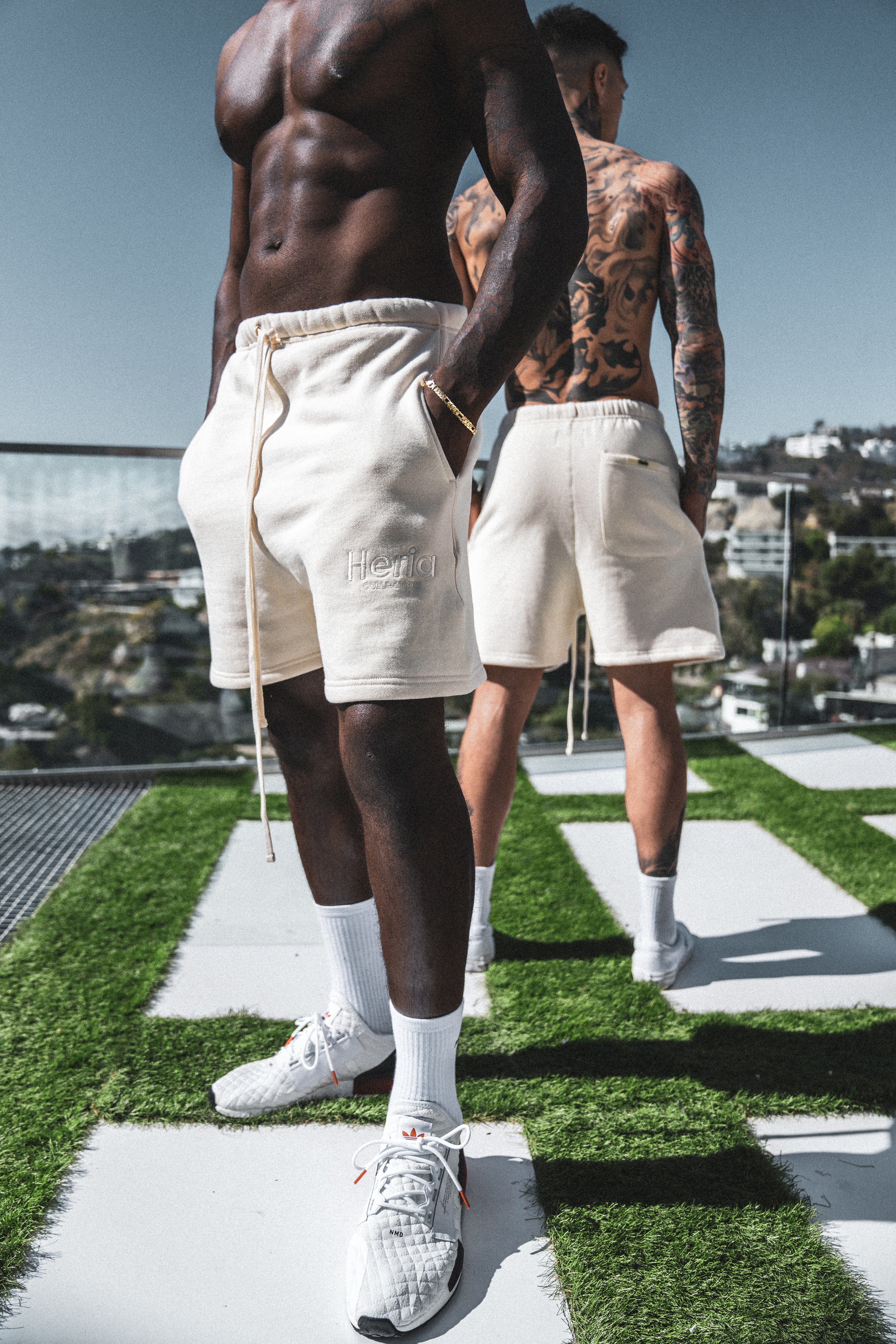 Two male models pose in cream Heria embroidered shorts under a sunny blue sky.