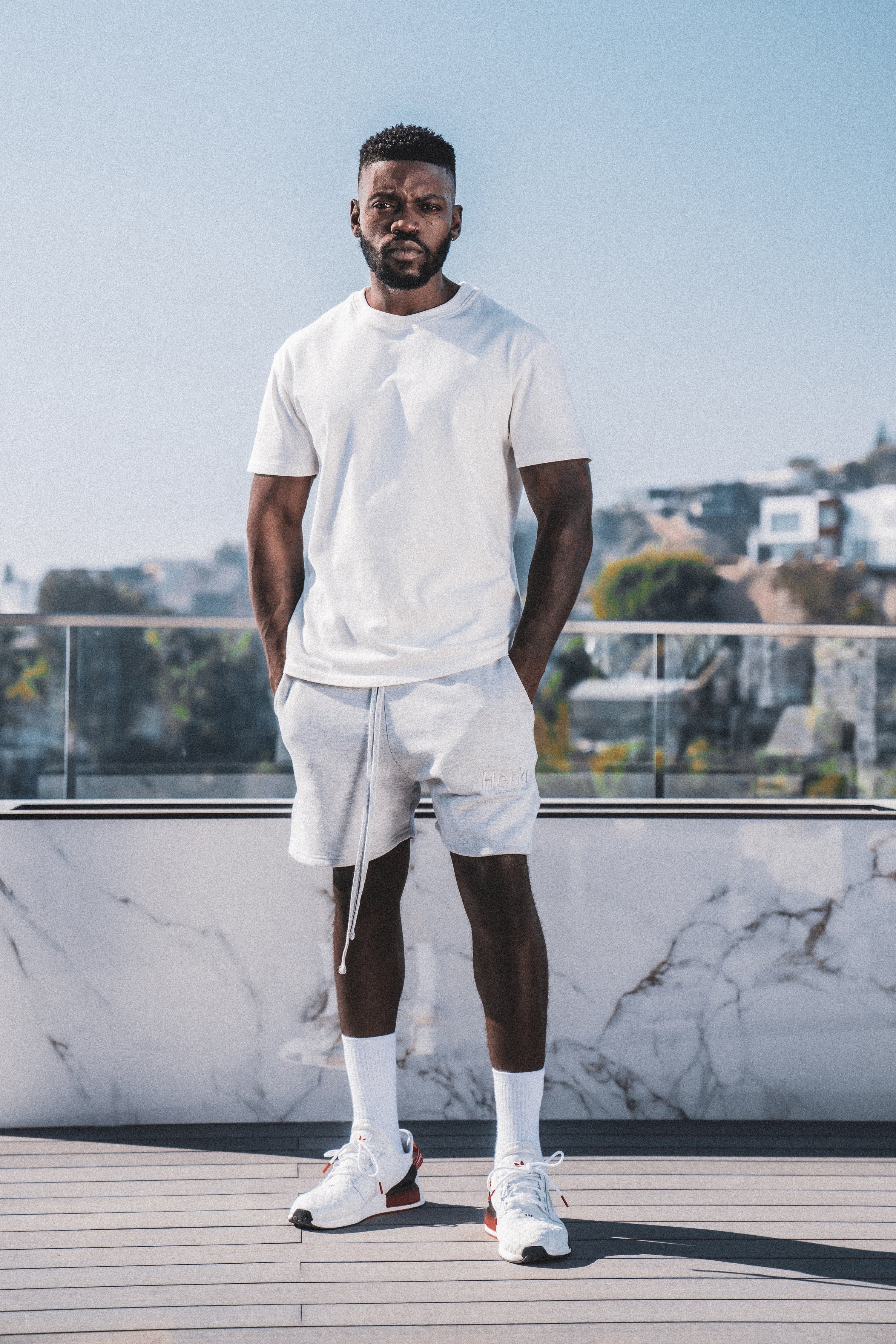 Male model posing under a sunny blue sky, wearing a white Heria t-shirt, grey shorts, and white Heria socks.